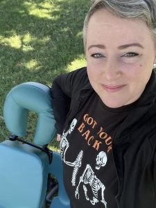 Vicki Hall smiling next to her massage chair, wearing a 'Got Your Back' shirt